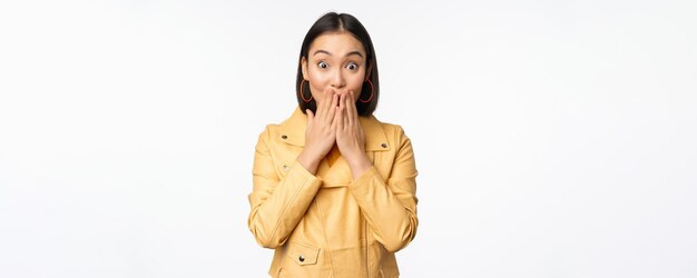 Portrait d'une fille asiatique excitée regardant avec intérêt la caméra étonnée de la réaction aux grandes nouvelles ou à l'annonce debout dans une veste jaune sur fond blanc