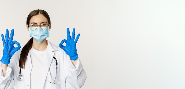 Photo gratuit portrait en gros plan d'une femme médecin médecin portant un masque facial contre le coronavirus montrant un bon signe d'approbation recommandant des éloges et comme qch bonne réputation sur fond blanc