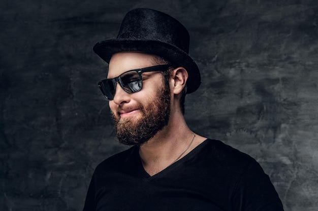 Photo gratuite portrait d'un homme barbu dans un t-shirt noir, un chapeau cylindrique et des lunettes de soleil sur fond gris.