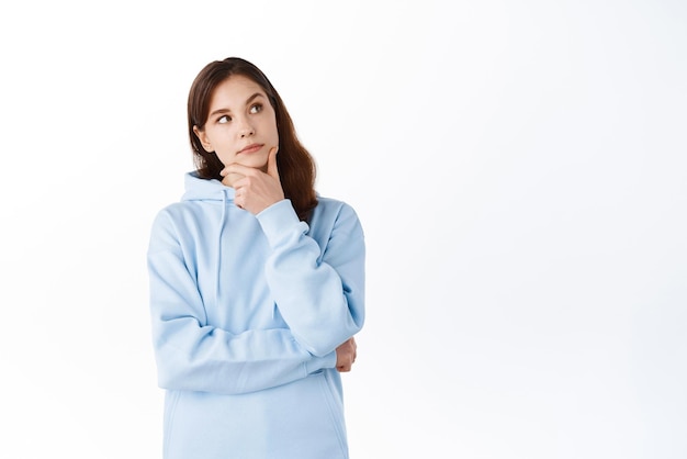 Photo gratuite portrait de jeune fille brune avec un visage pensif touchant le menton et regardant de côté le coin supérieur droit faisant des achats de choix et pensant debout sur fond blanc