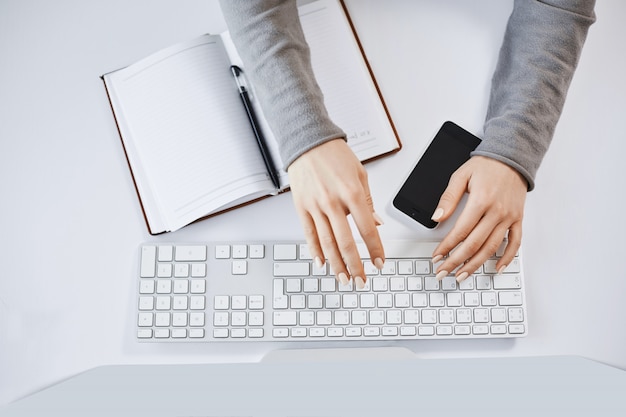 Portrait recadrée de mains de femme tapant sur le clavier et travaillant avec un ordinateur et des gadgets. Pigiste moderne concevant un nouveau projet pour l'entreprise, prenant des notes dans un ordinateur portable et un smartphone