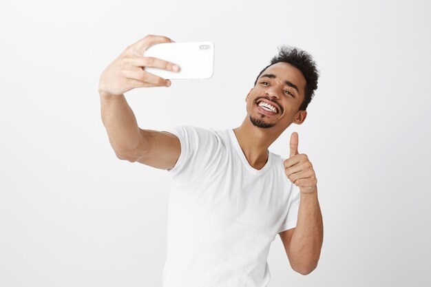 Sassy smiling guy afro-américain prenant selfie sur smartphone, montrant le geste de pouce en l'air