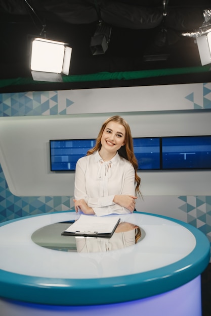 Photo gratuite télévision présente au studio se préparant pour une nouvelle diffusion. jeune fille souriante en chemise blanche assise à table.