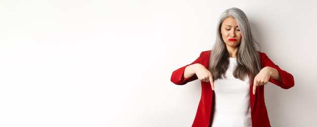 Photo gratuite triste et sombre femme âgée asiatique en blazer rouge boudant pointant du doigt vers le bas et regardant stan bouleversé