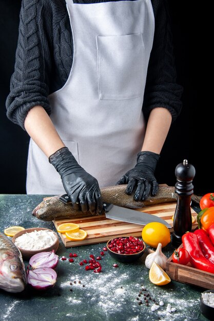 Vue de face chef avec des gants noirs hacher du poisson cru sur planche de bois moulin à poivre bol de farine graines de grenade dans un bol sur la table de la cuisine