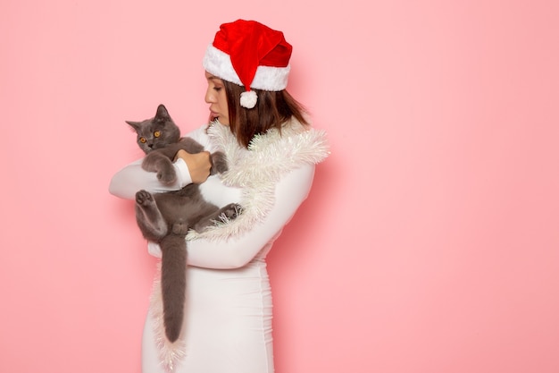 Photo gratuit vue de face d'une jeune femme en bonnet rouge tenant un joli chaton gris sur le mur rose