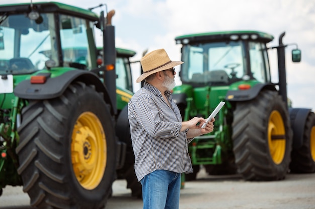 Photo agriculteur professionnel avec un tracteur moderne au travail avec tablette. semble confiant, couleurs estivales vives, soleil. agriculture, exposition, machines, production végétale. homme supérieur près de sa machine.