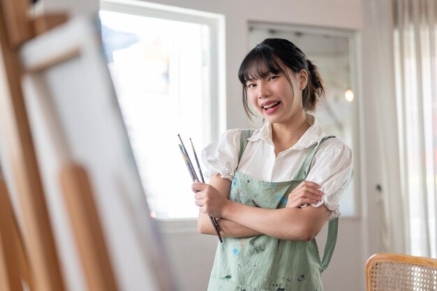 Photo une artiste féminine se tient les bras croisés et tient des pinceaux à la main dans son atelier