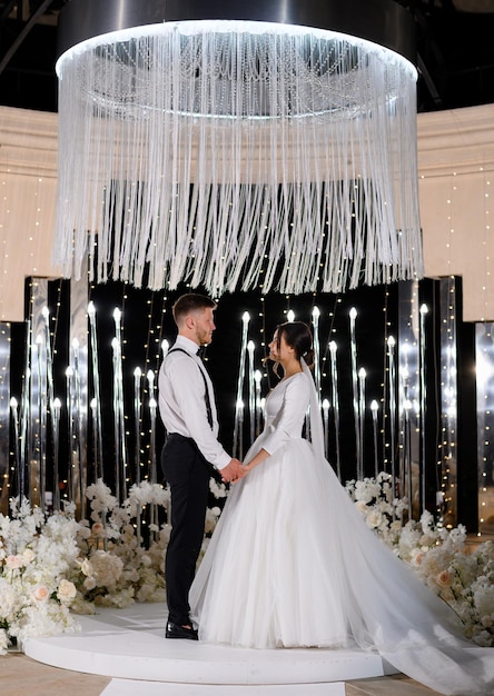 Beau couple de mariées aimantes debout sur un autel de luxe décoré de fleurs la nuit se regardant Femme élégante en robe longue bouffante avec bel homme Moment spécial dans la vie