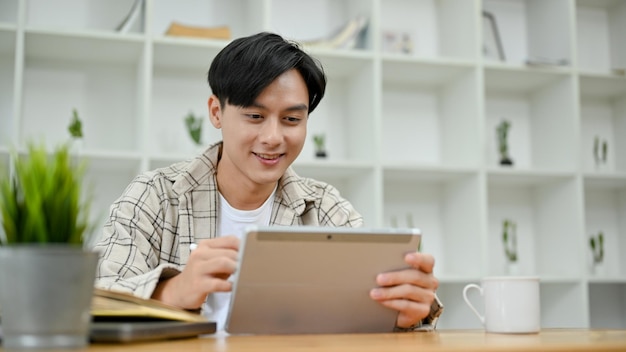 Bel homme asiatique en tenue causale à l'aide d'un pavé tactile de tablette numérique à son bureau dans la salle de bureau