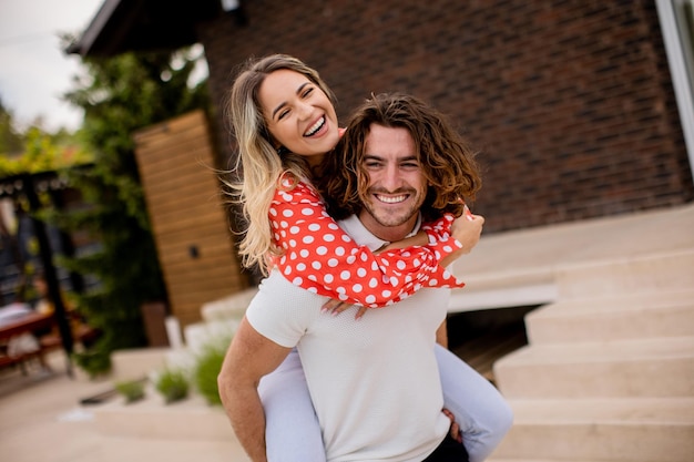 Bel homme aux cheveux longs portant la jeune femme sur le dos devant la maison en brique