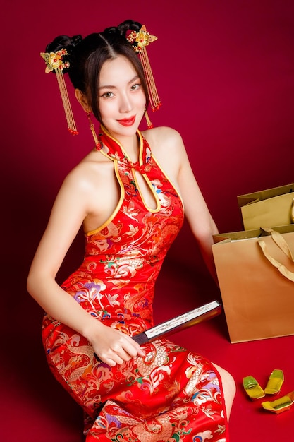 Belle femme asiatique avec une peau fraîche et propre portant une robe cheongsam tenant un ventilateur assis avec des sacs à provisions en or sur fond rouge Portrait de modèle féminin en studio Joyeux nouvel an chinois