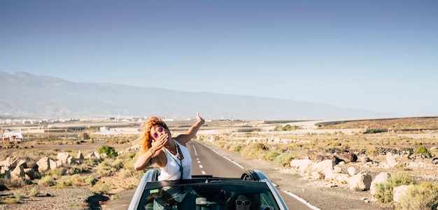 Photo belle jeune femme voyageuse envoie un baiser en restant hors du toit d'une voiture décapotable pendant que son amie conduit pour des voyages et des vacances à la campagne