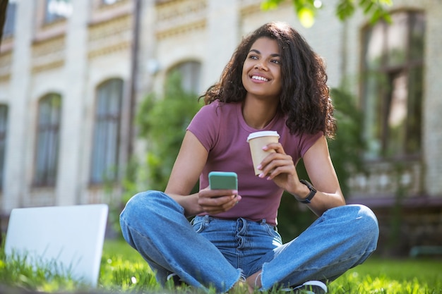 Café dans le parc. Une jolie fille en t-shirt rose assise sur l'herbe et prenant un café