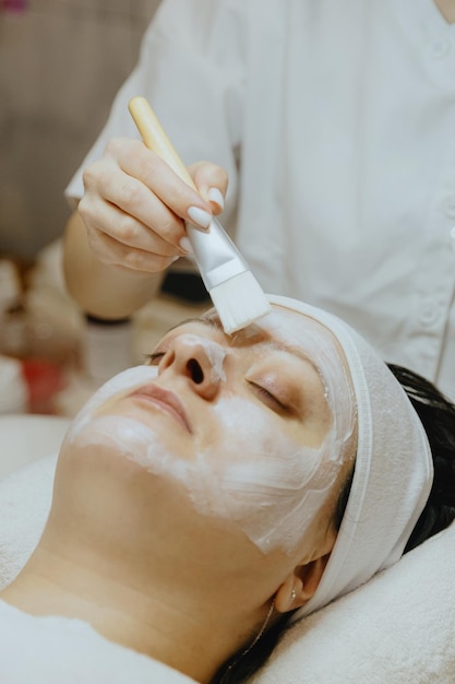 Une cosmétologue applique un masque de crème avec un pinceau au visage d'une femme