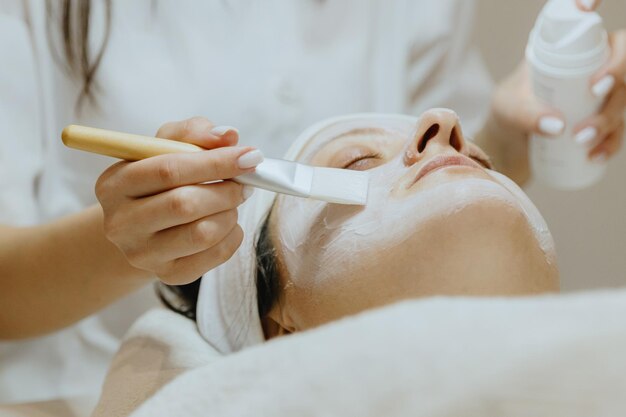 Une cosmétologue applique un masque de crème avec un pinceau au visage d'une femme