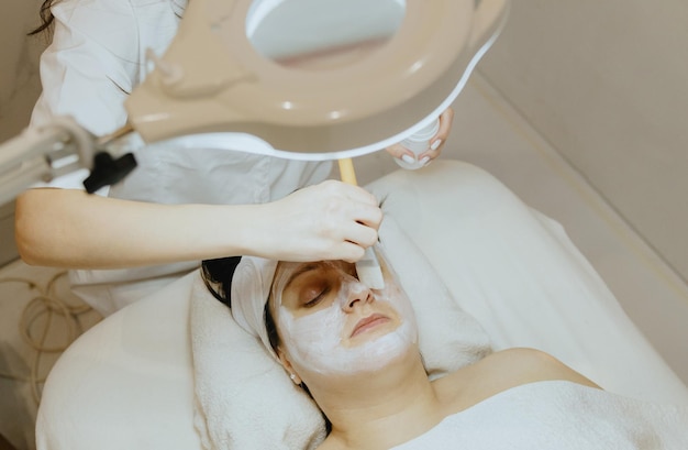 Une cosmétologue applique un masque de crème avec un pinceau au visage d'une femme