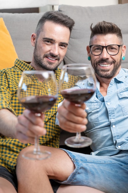 Couple gay amoureux assis à la maison sur le sol et portant un toast avec du vin rouge