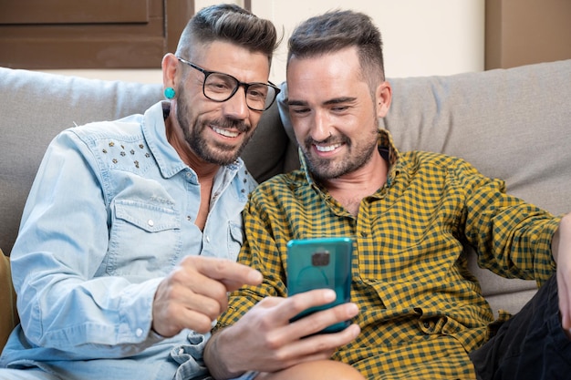 Couple gay assis dans le canapé à la maison faisant un appel vidéo sur téléphone mobile