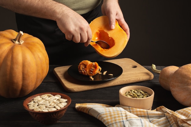 Le cuisinier sépare le grain de la citrouille coupée en deux. Sur une table en bois noire se trouvent des citrouilles entières de différentes tailles. Graines dans deux bols.