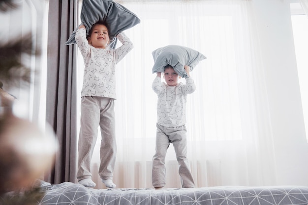 Photo les enfants jouent dans le lit des parents, les enfants se réveillent dans une chambre blanche ensoleillée, les garçons et les filles jouent en pyjama identique, le matin en famille.