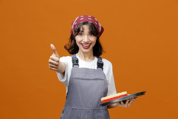 Excitée jeune femme nettoyante portant un uniforme et un bandana tenant un plateau avec une éponge dedans regardant la caméra montrant le pouce vers le haut isolé sur fond orange