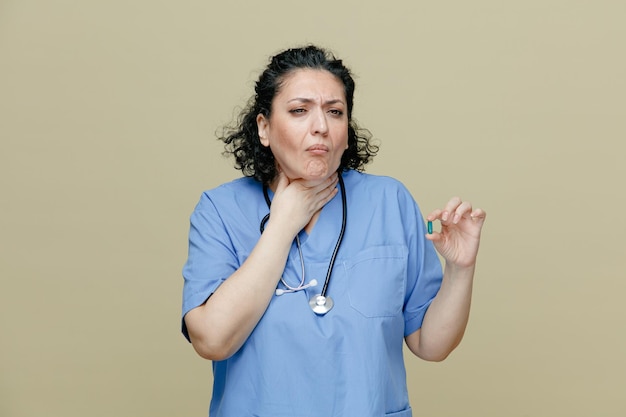 Femme médecin d'âge moyen douloureuse portant l'uniforme et le stéthoscope autour du cou montrant la capsule gardant la main sur le cou regardant le côté isolé sur fond olive