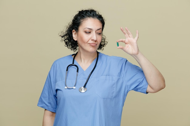 Femme médecin d'âge moyen incertaine portant un uniforme et un stéthoscope autour du cou montrant une capsule la regardant isolée sur fond olive