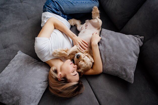 fille blonde en jeans est allongée sur le dos sur un canapé gris avec un chien