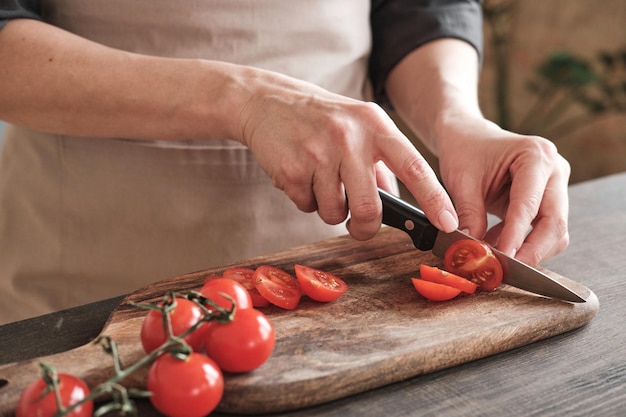 Gros plan sur des mains féminines coupant en tranches des tomates cerises avec un couteau sur une planche de bois faisant cuire une salade de légumes