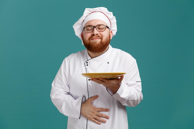 Heureux jeune chef masculin portant des lunettes uniformes et une casquette tenant une assiette vide montrant un geste affamé avec les yeux fermés isolé sur fond bleu