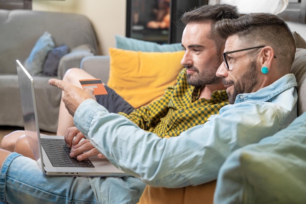 Heureux jeune couple gay utilisant un ordinateur portable assis sur un canapé à la maison faisant des achats en ligne avec une carte de crédit