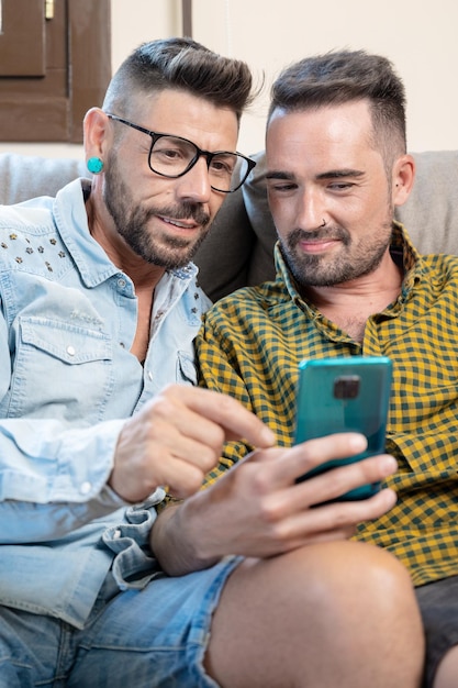 Heureux jeune couple gay utilisant un téléphone portable assis sur un canapé dans le salon