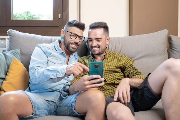 Heureux jeune couple gay utilisant un téléphone portable assis sur un canapé dans le salon