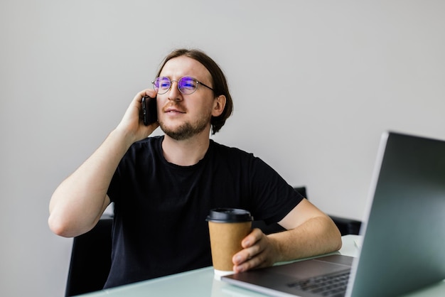 Heureux jeune homme parlant au téléphone portable et utilisant un ordinateur portable au bureau