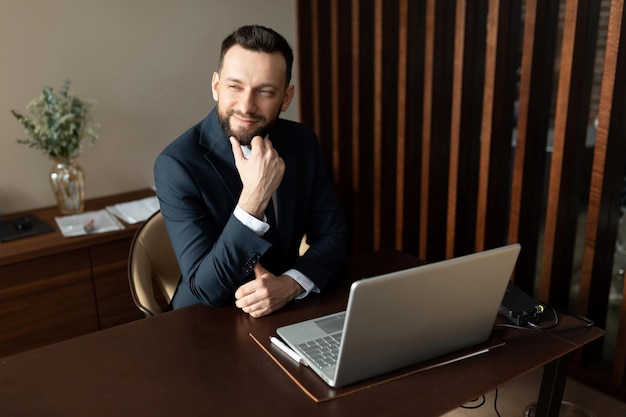 Homme d'affaires prospère sur le lieu de travail derrière un ordinateur portable en costume bleu regarde la caméra avec un