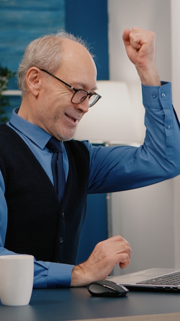 Un homme âgé excité se sent extatique en lisant d'excellentes nouvelles en ligne sur un ordinateur portable travaillant à domicile