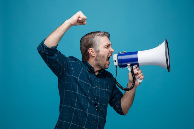 Homme d'âge moyen aux cheveux gris en chemise de couleur foncée criant dans un mégaphone étant excité et en colère serrant le poing debout sur fond bleu