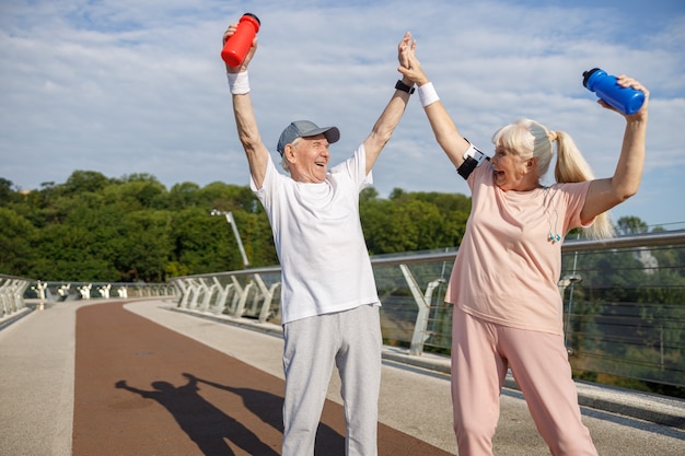 Photo un homme et une femme supérieurs gais soulèvent des bouteilles et joignent les mains sur la passerelle