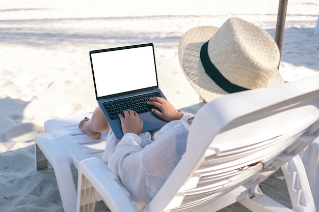 Image de maquette d'une femme à l'aide et en tapant sur un ordinateur portable avec écran de bureau vide tout en fixant sur une chaise de plage sur la plage