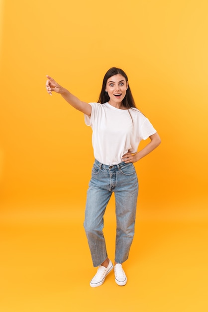 Image pleine longueur d'une femme brune excitée aux cheveux longs souriant et pointant du doigt sur le fond isolé sur un mur jaune