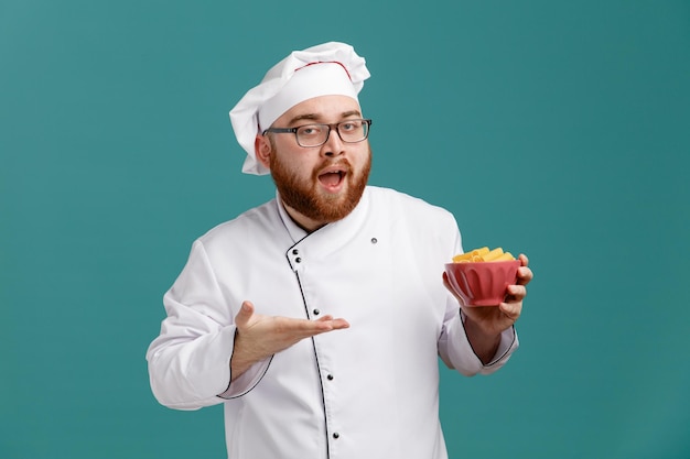 Impressionné jeune chef masculin portant des lunettes uniformes et une casquette tenant un bol de macaronis pointant avec la main dessus regardant la caméra isolée sur fond bleu