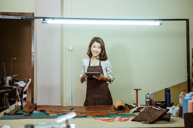 Photo jeune artisane asiatique souriant à la caméra tout en utilisant un tampon dans un atelier de cuir