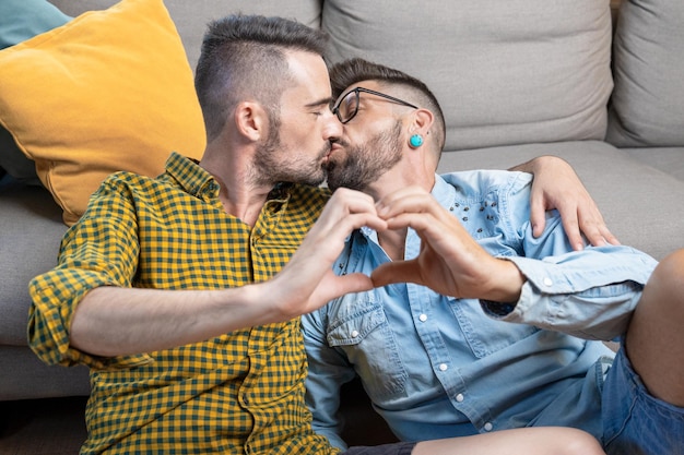 Jeune couple gay s'embrassant tout en faisant le symbole du cœur avec les mains à la maison