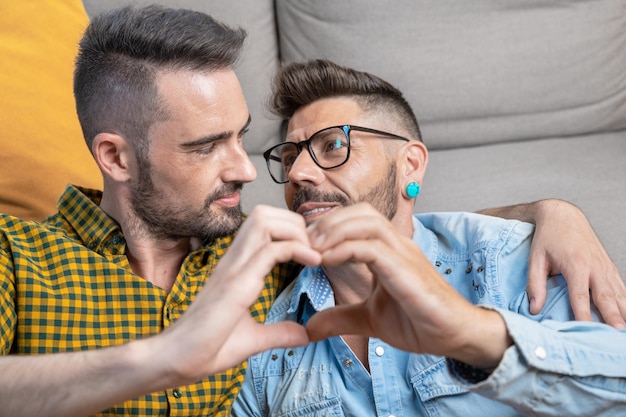 Jeune couple gay souriant confiant faisant le symbole du coeur avec les mains à la maison