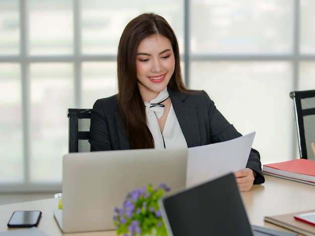 Jeune femme asiatique séduisante en affaires noires assises derrière une table avec un ordinateur et un ordinateur portable travaillant dans un bureau moderne avec un arrière-plan flou de fenêtres. Concept de style de vie de bureau moderne.