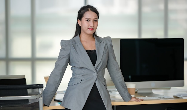 Jeune femme asiatique séduisante en costume d'affaires gris debout avec confiance dans un bureau moderne avec un arrière-plan flou de fenêtres. Concept de style de vie de bureau moderne.