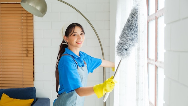 Photo jeune femme asiatique de service de nettoyage professionnel travaillant dans la maison la fille nettoie le rideau et la fenêtre avec un plumeau gris concept de service de nettoyage et de corvée professionnel