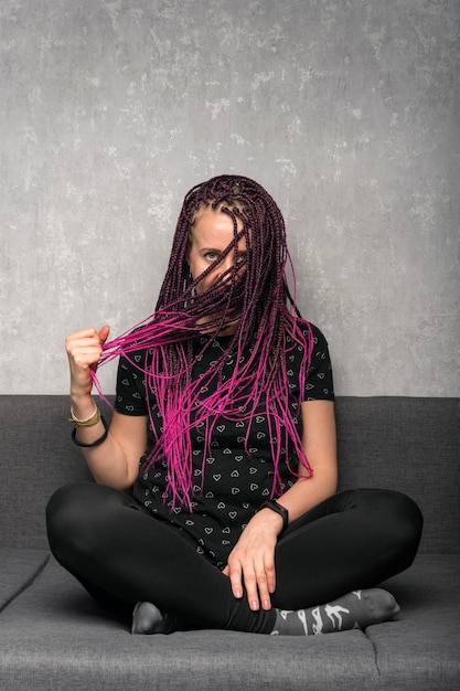 Photo jeune femme caucasienne avec des dreadlocks de couleur est assise sur le canapé et touche ses cheveux cadre vertical