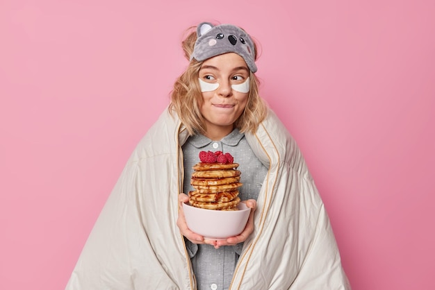 Une jeune femme européenne positive mord les lèvres tient des crêpes appétissantes regarde ailleurs enveloppé dans une couverture applique des patchs de beauté sous les yeux va manger un délicieux petit déjeuner isolé sur fond rose